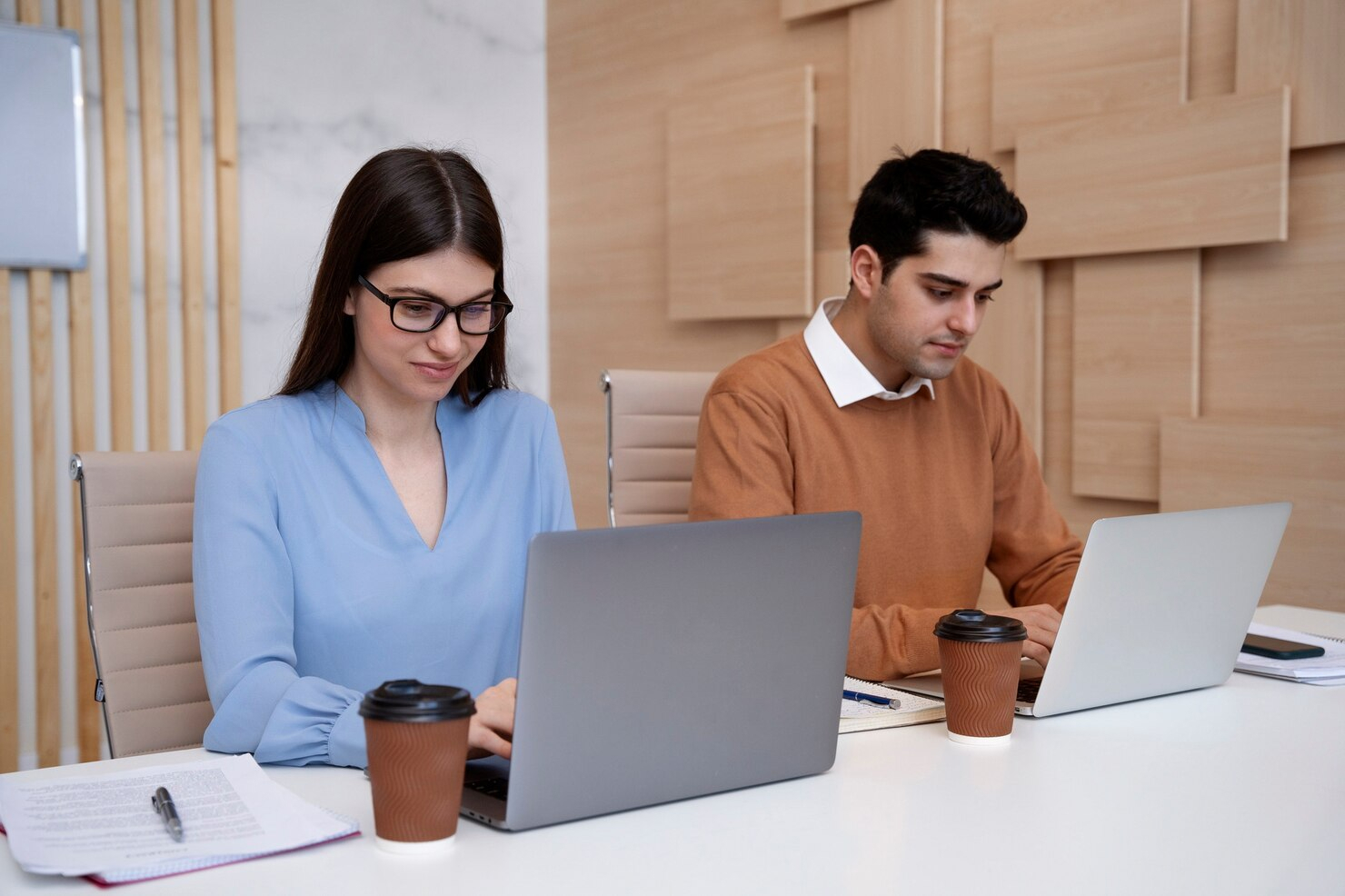 A young man and a young woman sitting in a modern office working independently on their laptops.